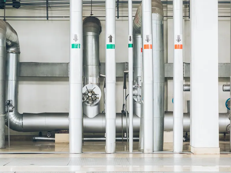 Insulated and steel service pipes stand in rows against a pale wall in a building plant room.