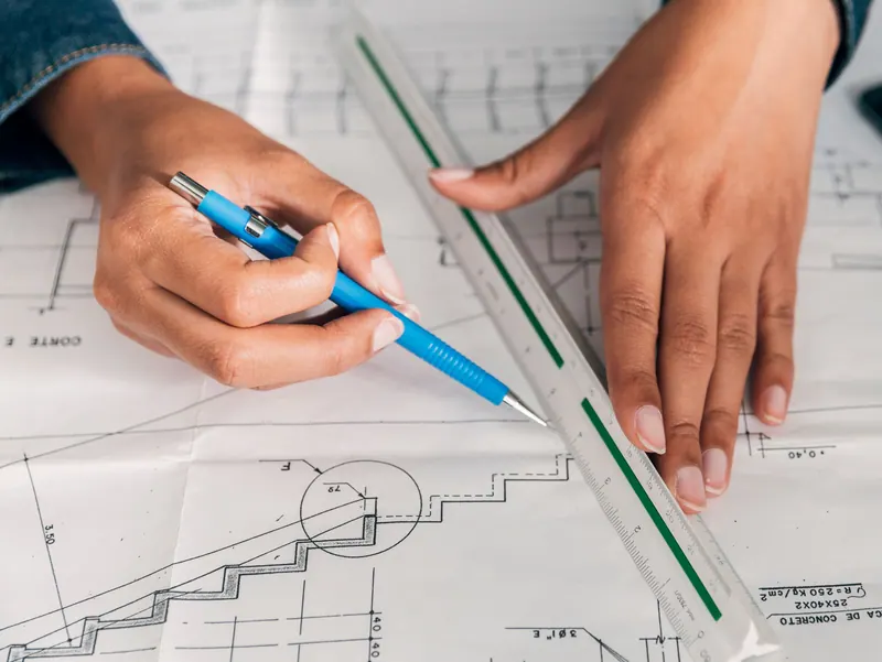 Hands hold a scale rule flat on a construction drawing while a blue pen marks a measurement.