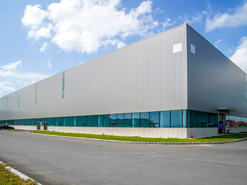 A long grey distribution warehouse with a glazed office strip along its base, under a light cloudy sky.